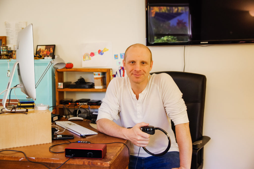 Hanuman Goleman is seen at his desk with podcasting equipment, desktop computer and TV in the background.